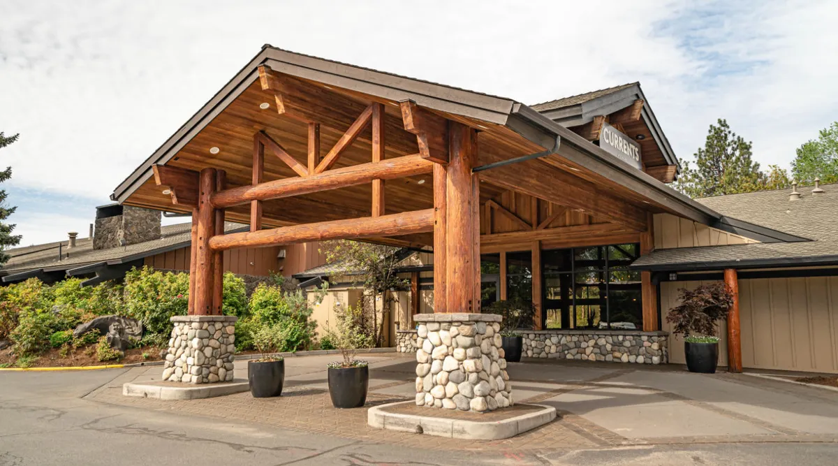 This exterior image features the main entrance of the property with impressive large wooden beams, stone columns, and a glass entryway surrounded by lush landscaped greenery. Guests are seen arriving, and small groups gather outdoors, suggesting a welcoming environment perfect for families and friends beginning their adventures.
