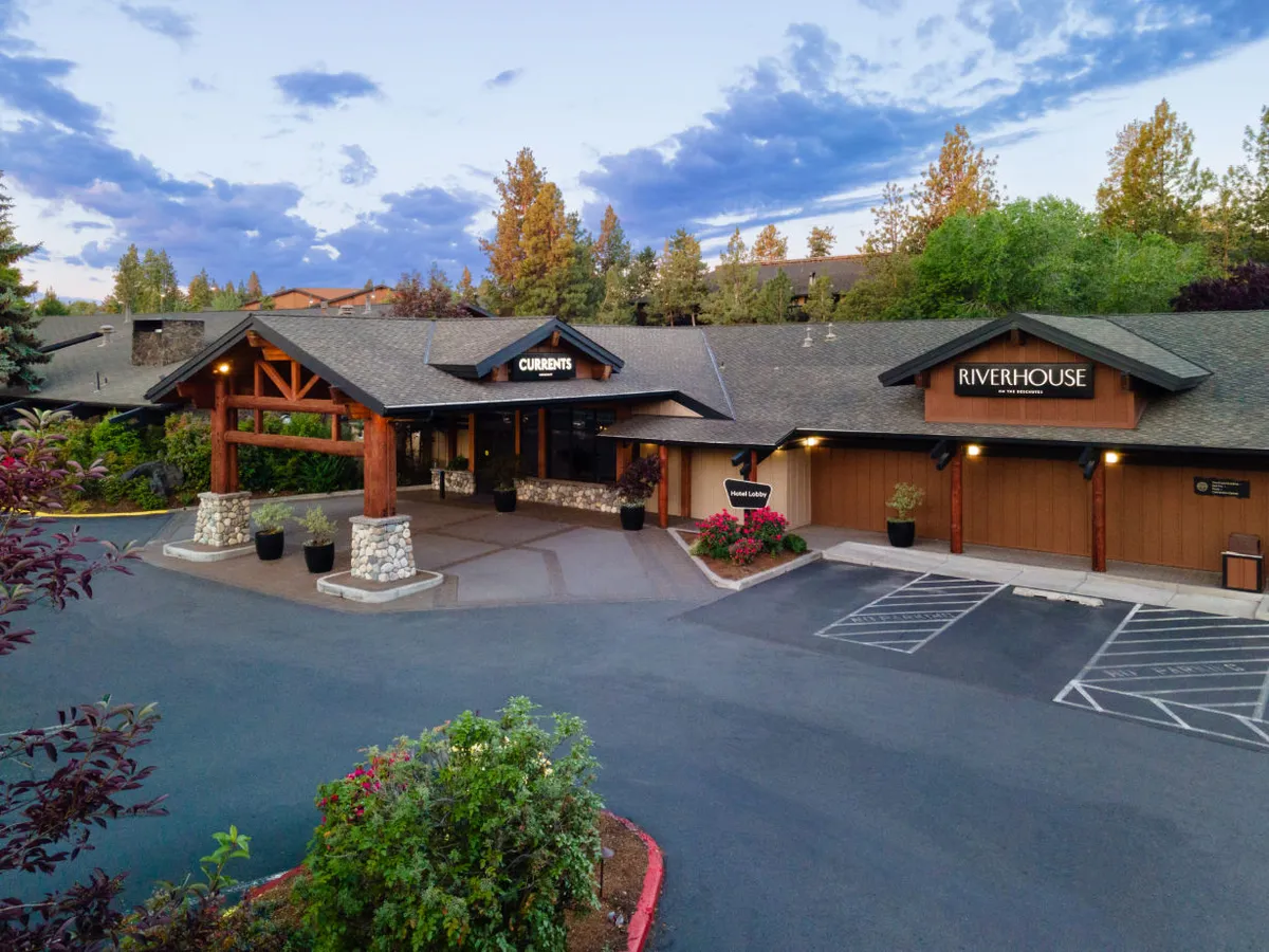 The exterior of the property featuring a wooden entrance, sturdy stone columns, and signage for 'Currents' and 'Riverhouse,' set amidst landscaped flower beds and bathed in serene, warm light. The image conveys a welcoming atmosphere for guests.