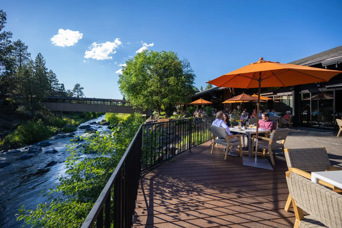 This image captures the outdoor riverside patio, where patrons are enjoying meals under bright orange umbrellas. The river is prominent in the background, creating a cheerful, relaxed ambiance. People are engaged in dining, socializing, and soaking up the scenic river views.