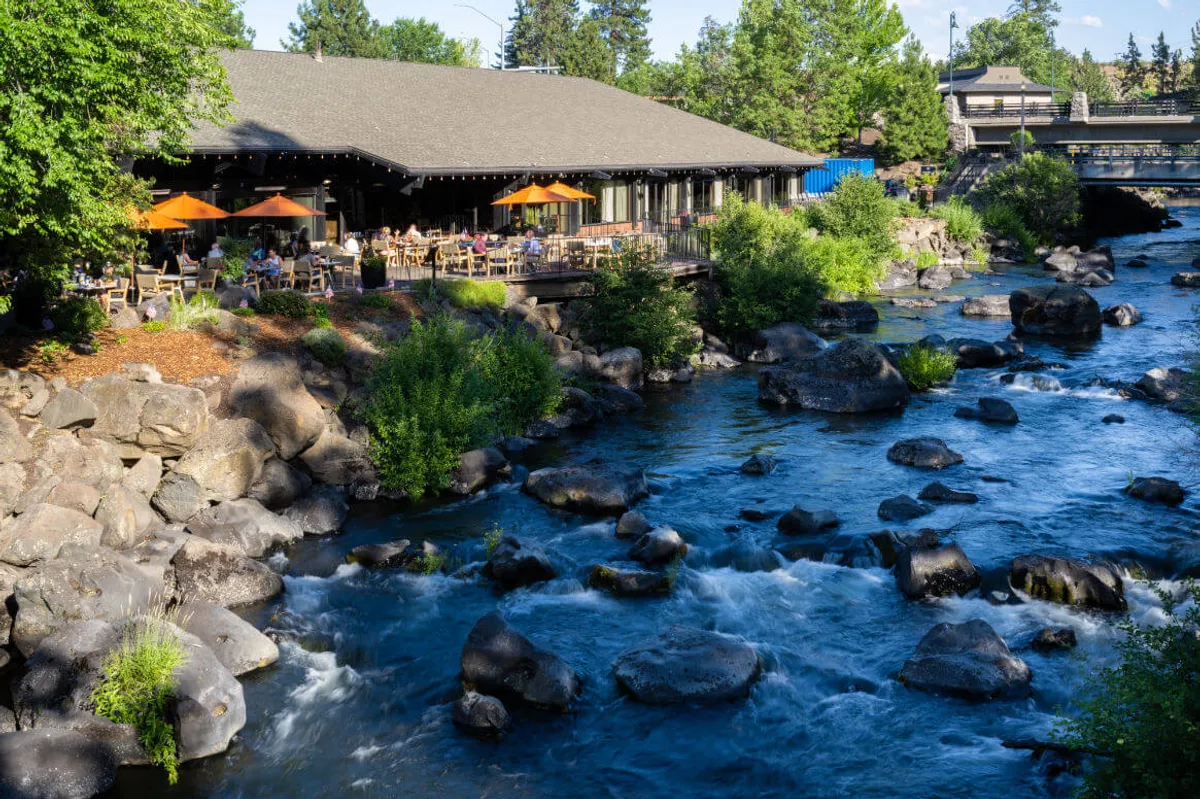 The image showcases the inviting outdoor restaurant patio of the Riverhouse Lodge overlooking a tranquil river, dotted with vivid orange umbrellas. Lush green foliage surrounds the scene, and large rocks line the water’s edge, creating a serene natural ambiance perfect for dining, relaxing, or gathering with friends.