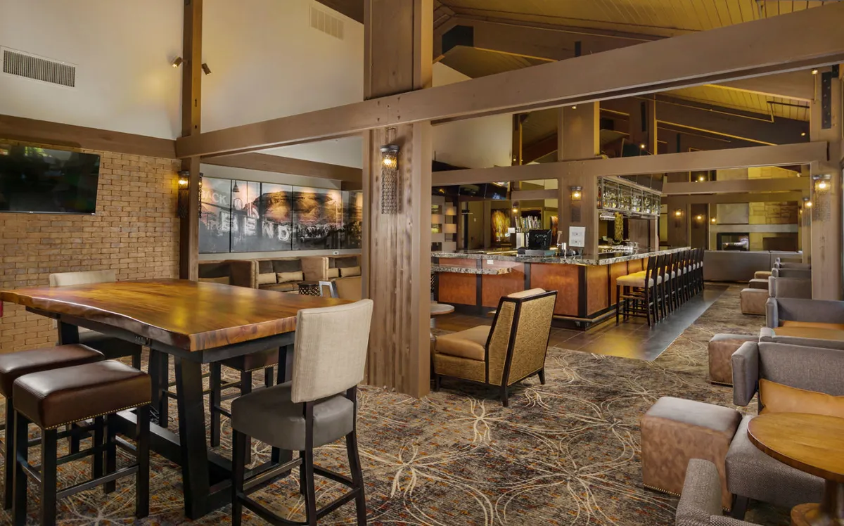 A spacious interior with a large wooden dining table surrounded by comfortable seating, positioned near a bar area with decorative wall art and warm lighting. The setting is casual yet sophisticated, suitable for meals, casual gatherings, and conversations.