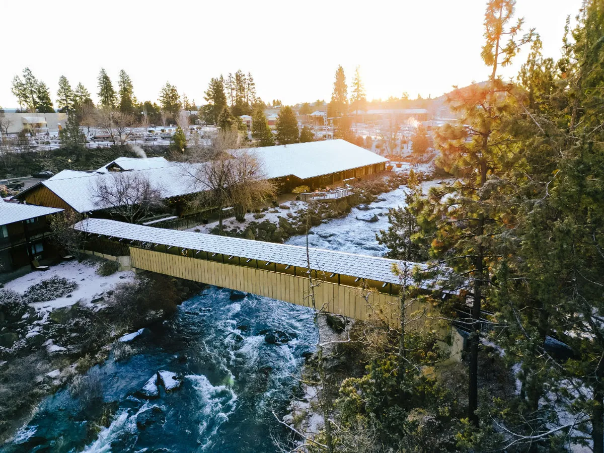 A peaceful scene showing a covered bridge over the river, surrounded by trees and snow-capped roofs in the distance. The setting is tranquil and scenic, inviting exploration and relaxation along the riverside.