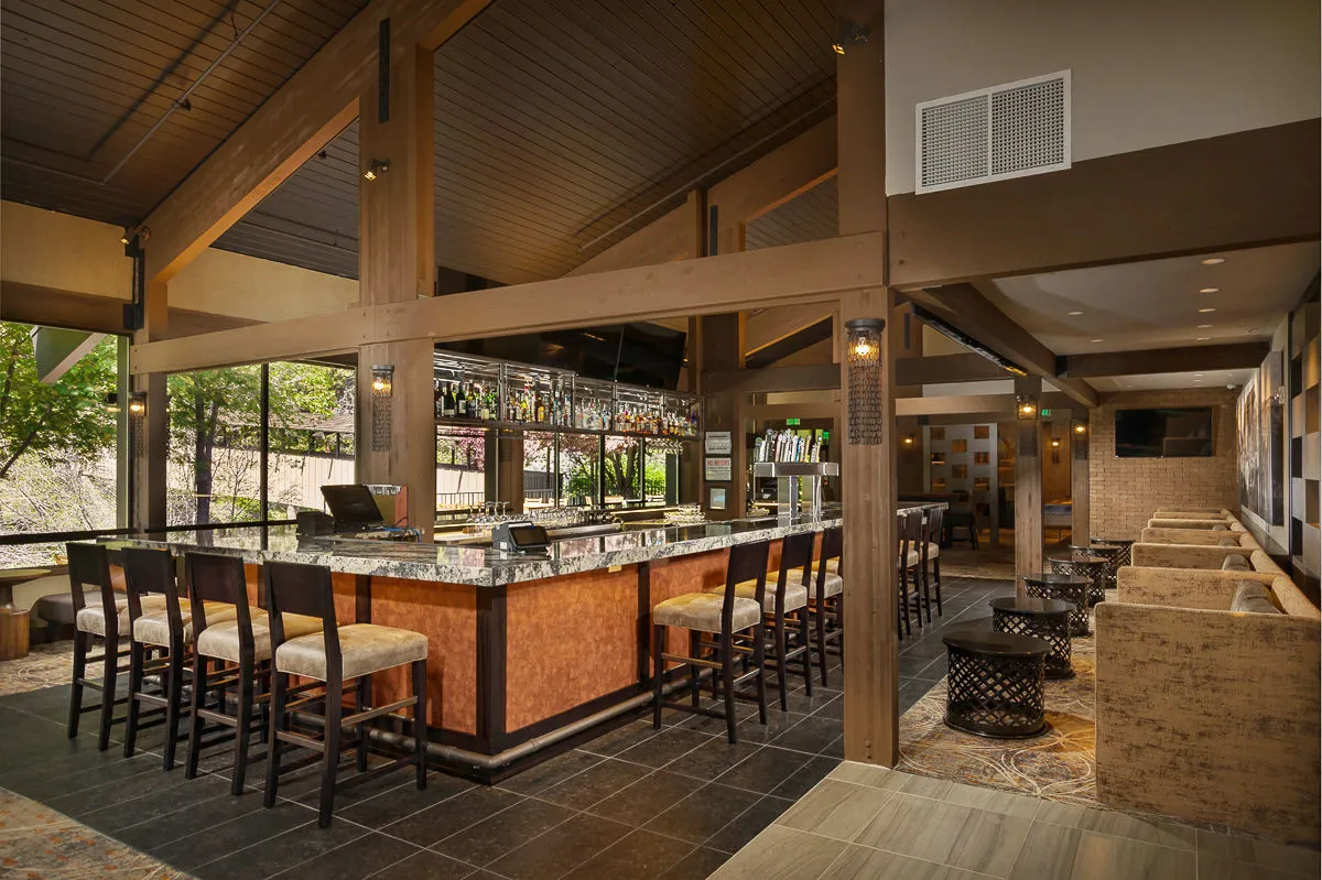 A stylish bar counter with elegant stools faces shelves lined with assorted bottles, while a comfortable sitting area invites guests to relax and socialize. Ambient lighting and a modern design create a warm, inviting mood, perfect for evening gatherings or an intimate cocktail hour.