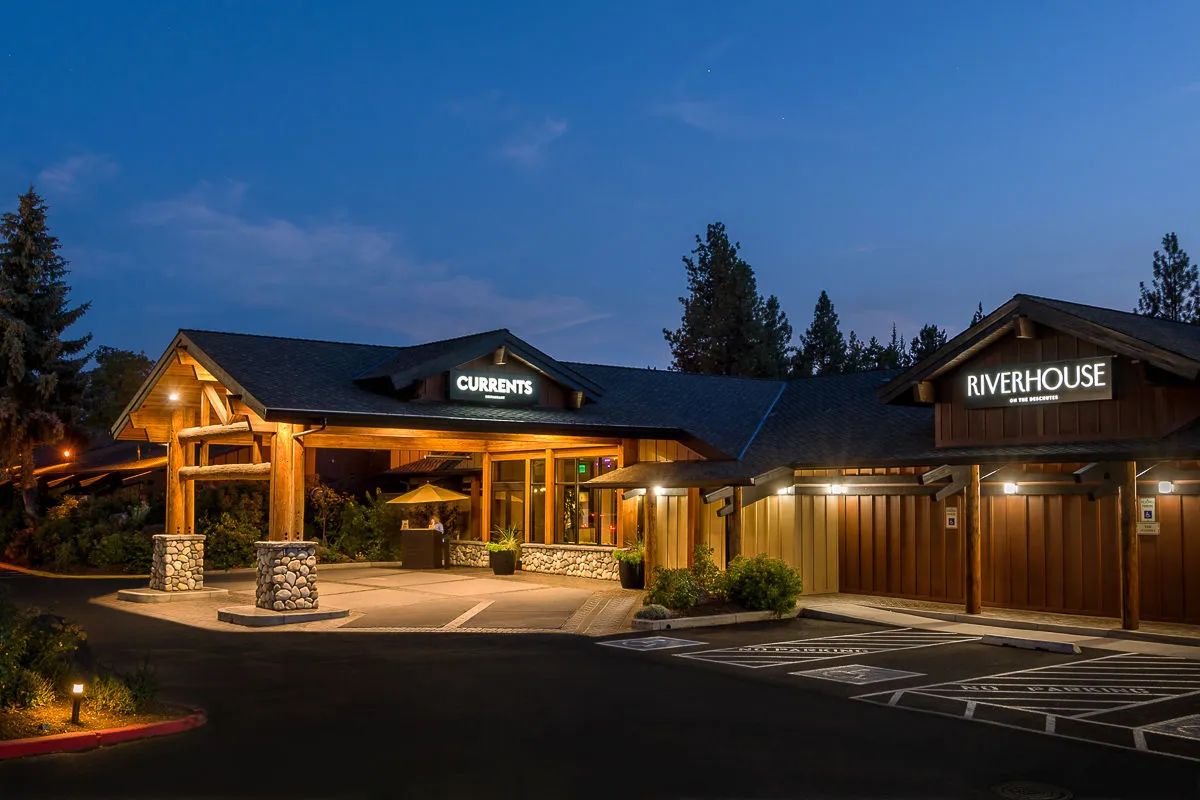 The exterior of the property features the entrance awning and signage for both CURRANTS and RIVERHOUSE. The landscaped area around the entrance further emphasizes the welcoming environment, with hints of dining and event spaces visible.