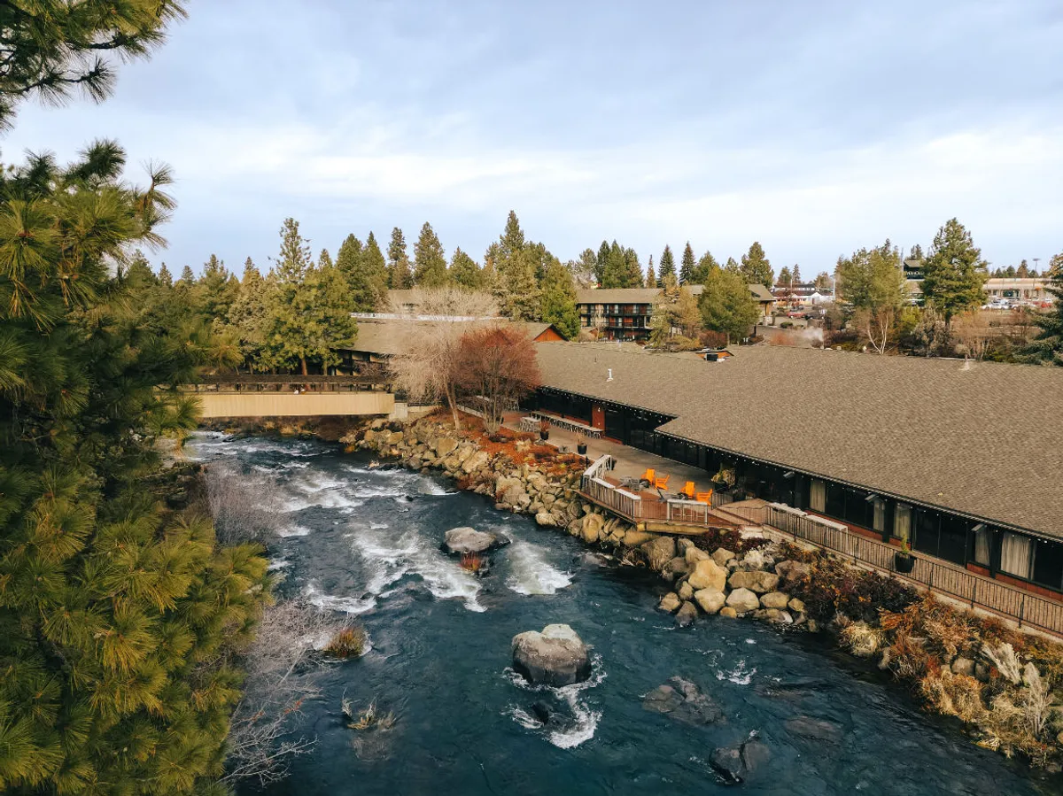 An inviting outdoor patio area overlooking the river, with tables and chairs set under the trees. The building exterior and surrounding nature offer a tranquil setting for relaxation or dining al fresco, with views that change by suite.