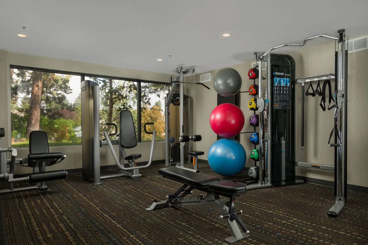 A bright, spacious fitness center equipped with modern exercise machines and colorful exercise balls, all set beside large windows that let in natural light and offer views of the outdoors. The room feels energetic and welcoming, encouraging wellness and activity.