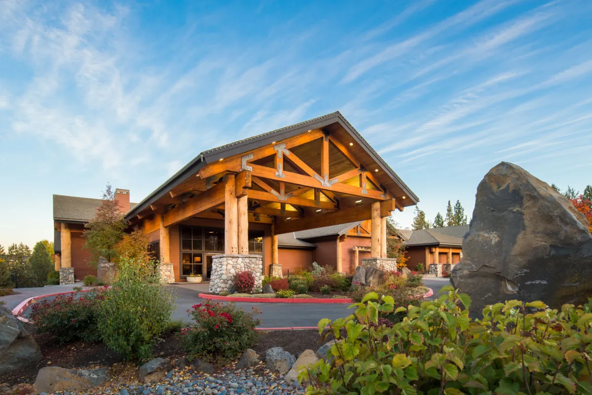 An exterior scene highlighting the property's rustic architecture with large wooden beams and a stone foundation, set against a vibrant blue sky. Landscaped gardens frame the entrance, while guests check in or gather for photos, conveying a lively and inviting atmosphere.