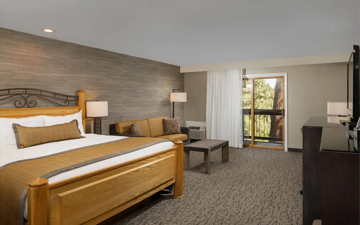A warmly lit suite interior showing a wooden king bed with crisp white linens, accompanied by a plush sofa seating area set near a large window that fills the room with natural light. The decor is unique and welcoming, featuring soft textures and warm earth tones.